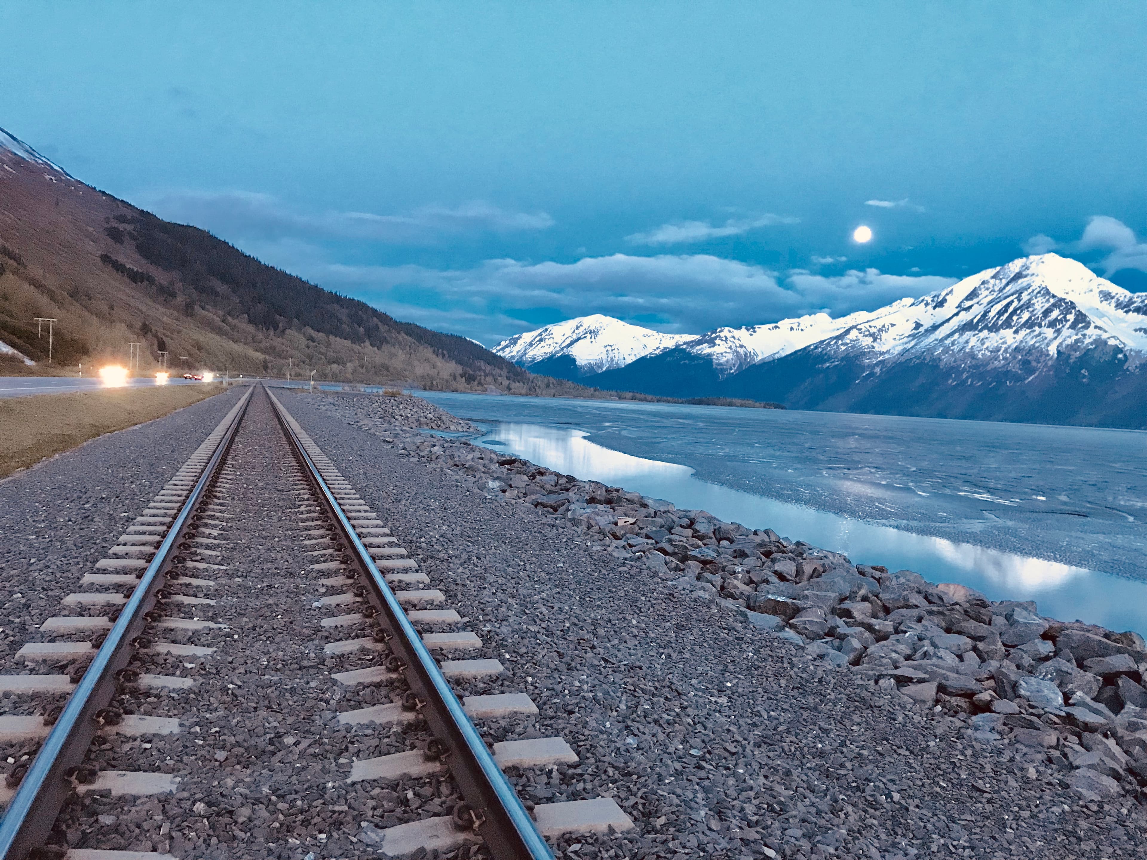 Railway tracks alongside frozen lake with snow-capped mountains at twilight